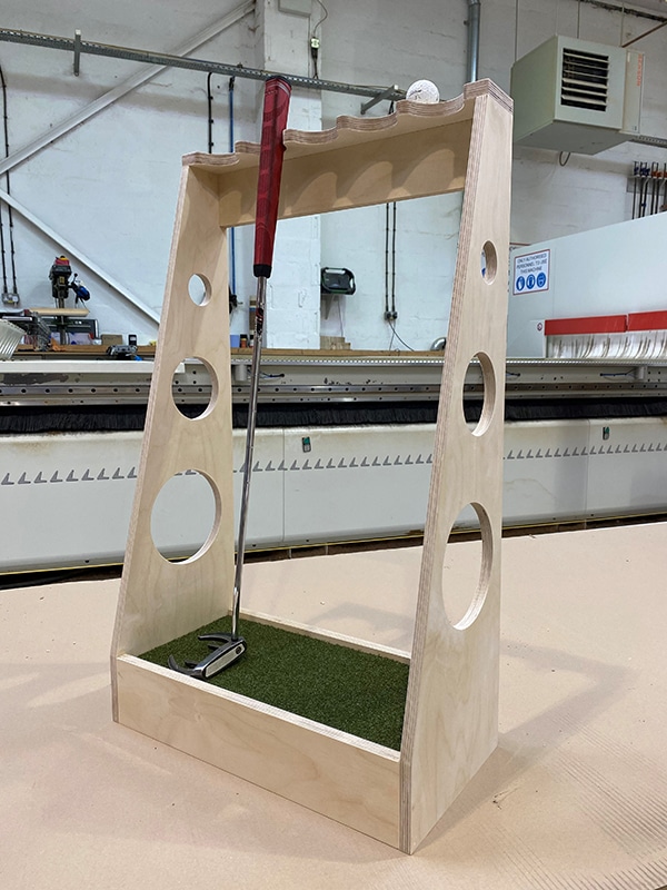 A staged photo showing bespoke CNC birch plywood putter for use on a back garden putting green. The photo was taken in a workshop and includes a putter and golf ball for effect